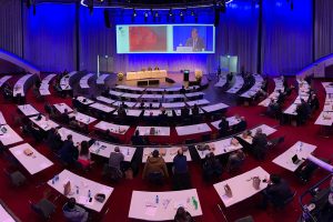 Vue plongeante d'un grand auditorium rempli lors d'une conférence, mettant en avant une installation complète par un intégrateur de solutions audiovisuelles. Un orateur en direct est sonorisé et capté en vidéo, tandis qu'un écran géant, partie intégrante des solutions d'affichage avancées, diffuse sa présentation multimédia. L'infrastructure intégrée assure la communication et la diffusion