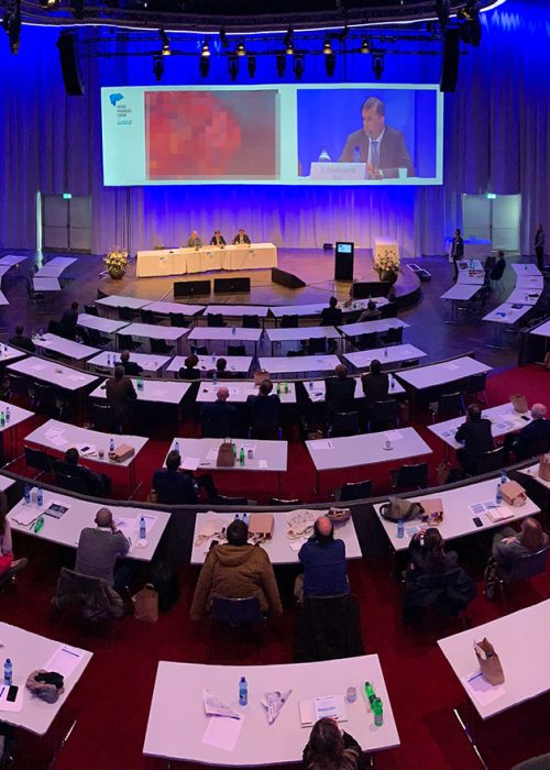 Vue plongeante d'un grand auditorium rempli lors d'une conférence, mettant en avant une installation complète par un intégrateur de solutions audiovisuelles. Un orateur en direct est sonorisé et capté en vidéo, tandis qu'un écran géant, partie intégrante des solutions d'affichage avancées, diffuse sa présentation multimédia. L'infrastructure intégrée assure la communication et la diffusion